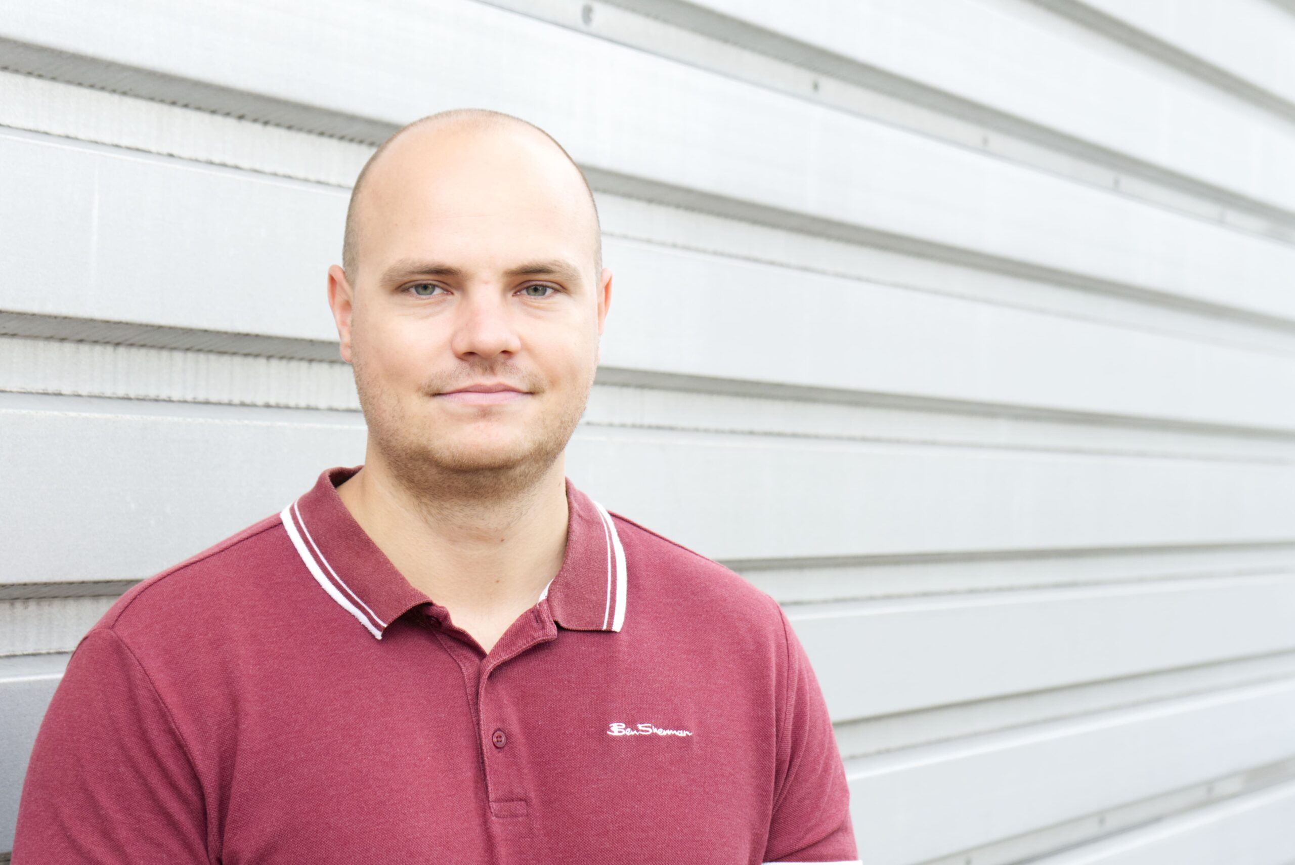 Ein junger Mann mit rasiertem Kopf und bordeauxrotem Poloshirt steht vor einer hellgrauen Metallwand und blickt freundlich in die Kamera.